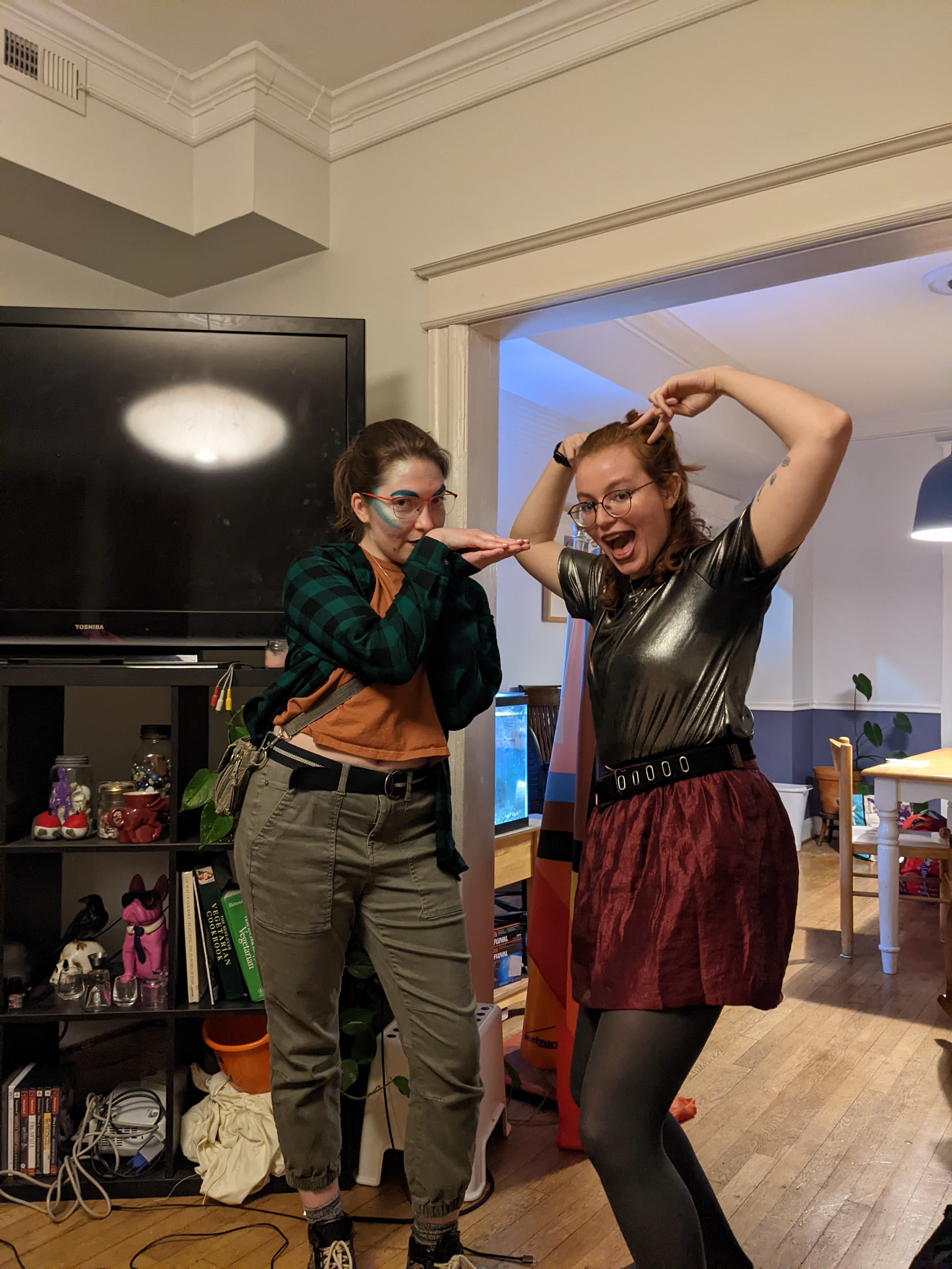 Two young adults in going out outfits posing in front of a shelf and tv.